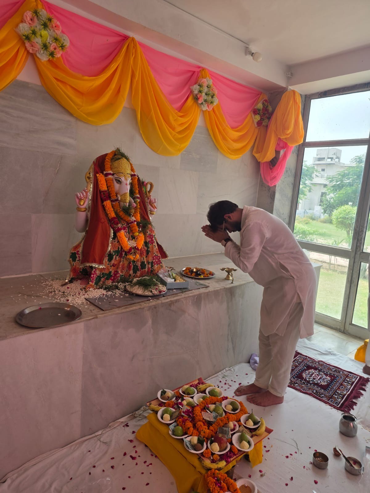 A man bowing with folded hands before a garlanded murti at Hansi Gita Bhawan, a thali of lamps and offerings on the step in front of him